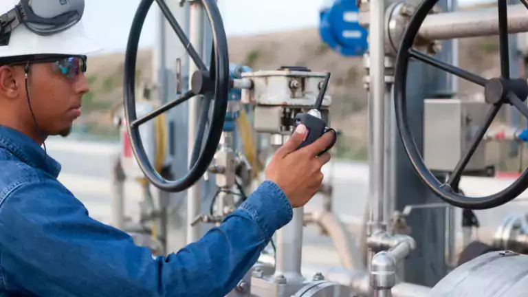 Industrial worker wearing safety gear operates a valve at a facility, holding a walkie-talkie and inspecting metal pipes and machinery outdoors.