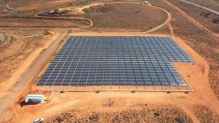 Aerial view of a large solar farm with many rows of solar panels installed on flat, dry land surrounded by dirt roads and sparse vegetation.