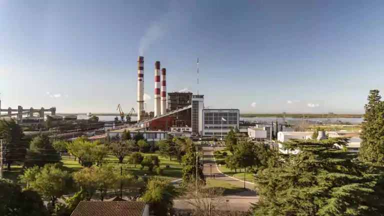 A large power plant with three tall smokestacks emitting light smoke, surrounded by green trees and a landscaped area, with a river and industrial cranes in the background under a clear blue sky.
