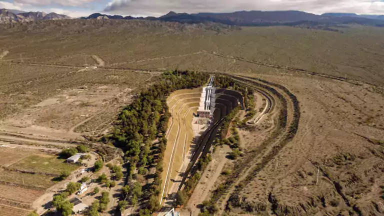 Aerial view of a large outdoor amphitheater surrounded by trees and desert landscape, with mountains and cloudy sky in the background.