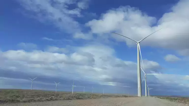 A row of wind turbines stands along a dirt road in a flat, open landscape under a blue sky with scattered clouds, generating renewable energy.
