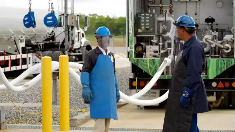 Two workers wearing protective gear, including helmets, gloves, aprons, and face shields, stand near a tanker truck at an industrial site. Hoses are connected to the truck, and safety equipment is visible in the background.