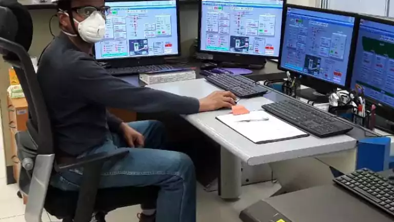 A person wearing a hard hat, safety glasses, and a face mask sits at a control room desk with four computer monitors displaying industrial system data.