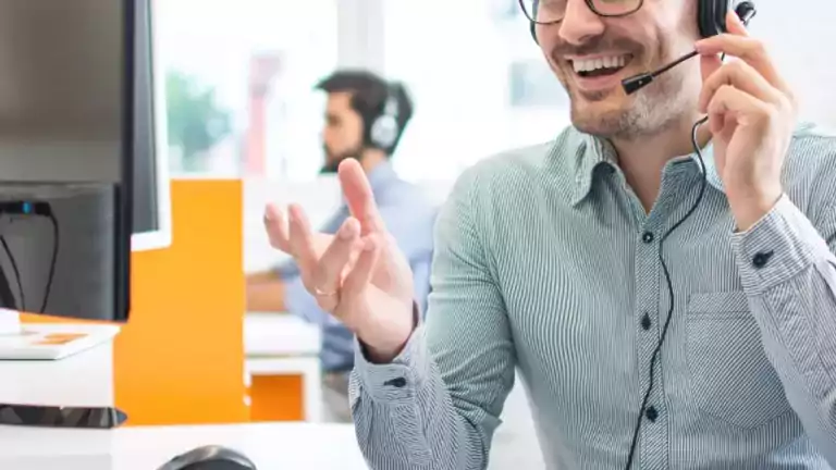 Smiling man wearing glasses and a headset sits at a desk with a computer, engaging in a conversation in a bright office environment. Another person with a headset is visible in the background.