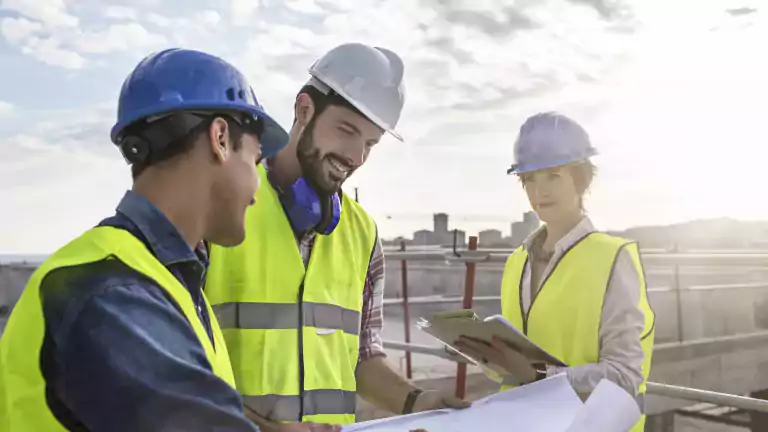 Three construction workers wearing safety vests and helmets are discussing blueprints at a construction site. One woman holds a clipboard while the two men review plans together under a partly cloudy sky.