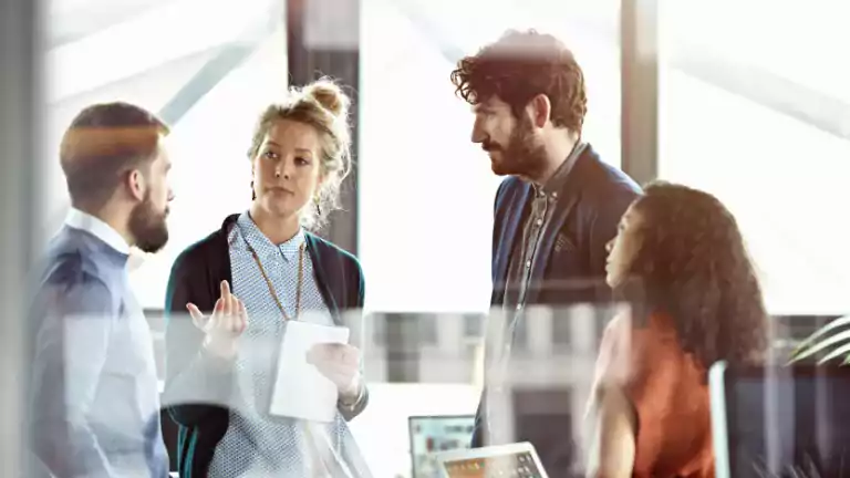 Four colleagues have a discussion in a modern office, standing together near large windows. One woman holds a tablet and gestures while speaking, as the others listen attentively. The scene suggests teamwork and collaboration in a professional workspace.