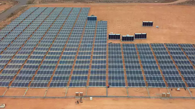 Aerial view of a large solar farm with rows of solar panels installed on a dry, sandy landscape, surrounded by barren land and dirt roads.