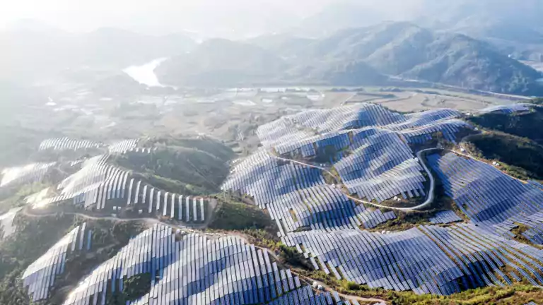Aerial view of a large solar farm with rows of solar panels covering rolling hills and green landscape, surrounded by mountains and a river in the distance under a hazy sky.