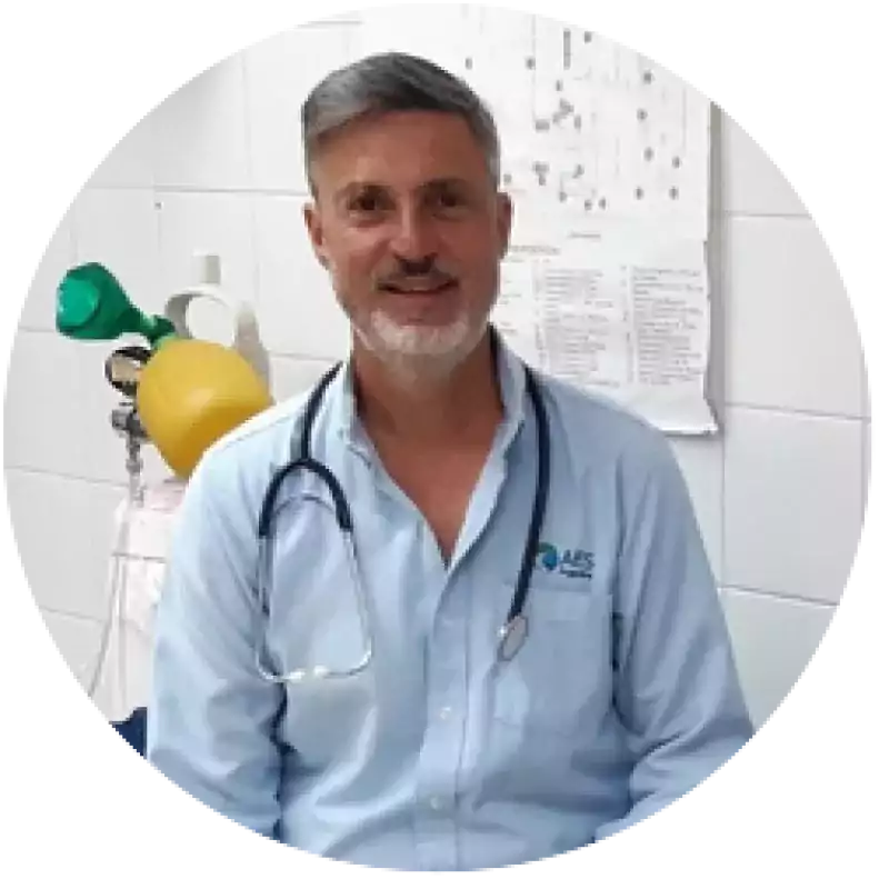 Male doctor with gray hair and beard wearing a light blue shirt and stethoscope, sitting in a medical office with medical equipment and charts in the background.
