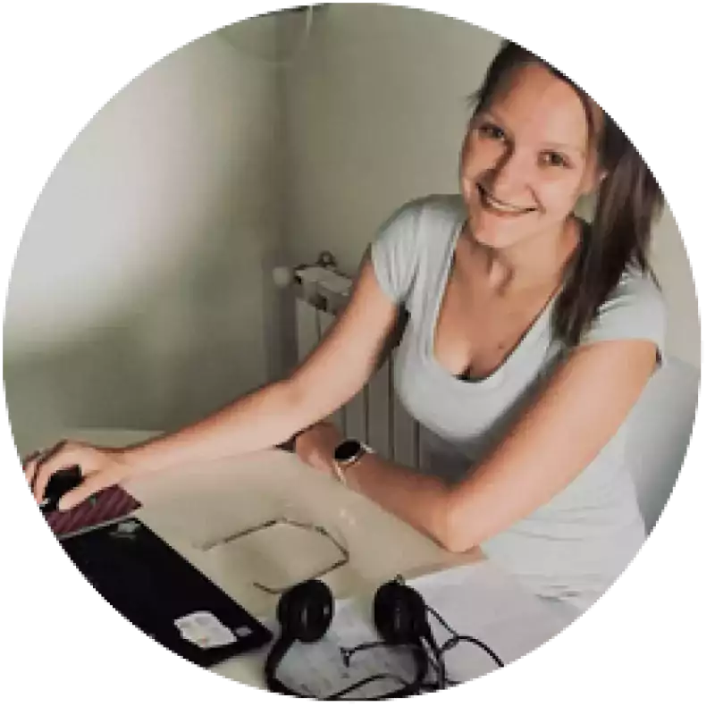 Smiling woman in a light gray shirt working at a desk with a laptop, headphones, and papers in a home office setting.