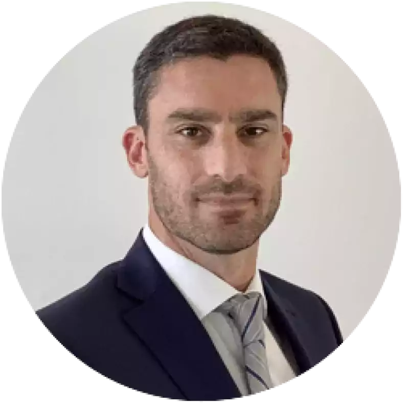 Man with short dark hair and trimmed beard wearing a dark suit, white shirt, and striped tie, standing in front of a plain light background.