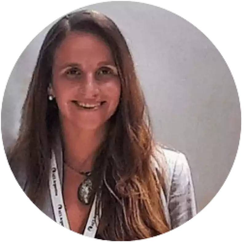 Smiling woman with long brown hair wearing a light-colored blazer, a necklace with a large pendant, and a lanyard, standing against a plain background.