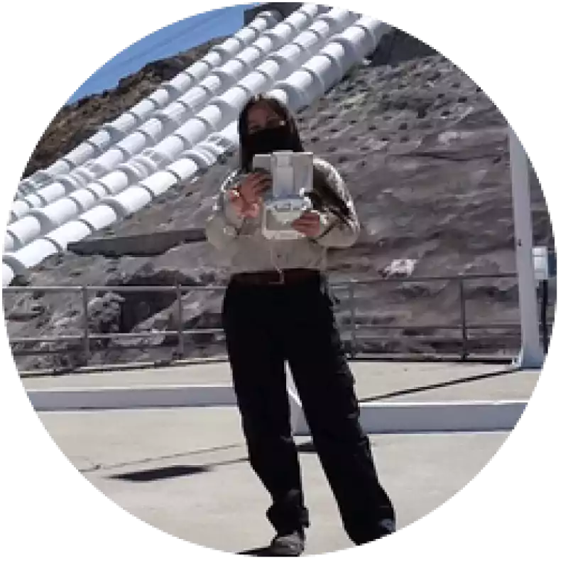 Person wearing a mask and holding a drone controller stands outdoors near a large industrial structure with pipes and rocky terrain in the background.