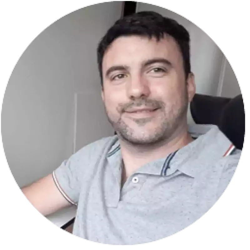 Man with short dark hair and trimmed beard wearing a light gray polo shirt, smiling while sitting in an office chair near a white wall.