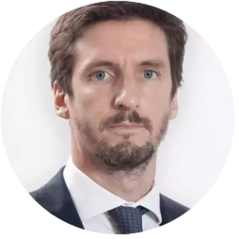 Portrait of a man with short brown hair, trimmed beard, and blue eyes, wearing a dark suit, white shirt, and patterned tie, against a plain white background.