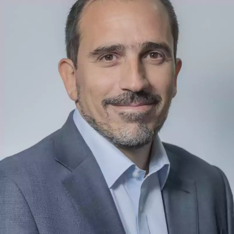 Portrait of a middle-aged man with short dark hair, beard and mustache, wearing a light blue shirt and a gray suit jacket, smiling softly against a plain light background.