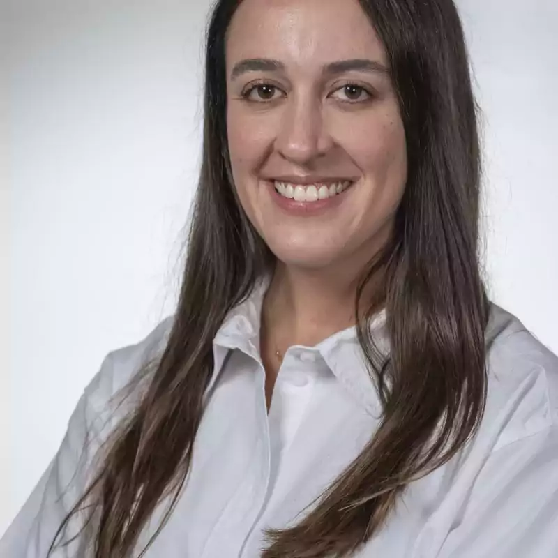Smiling woman with long brown hair wearing a white button-up shirt, standing with arms crossed against a plain light background.