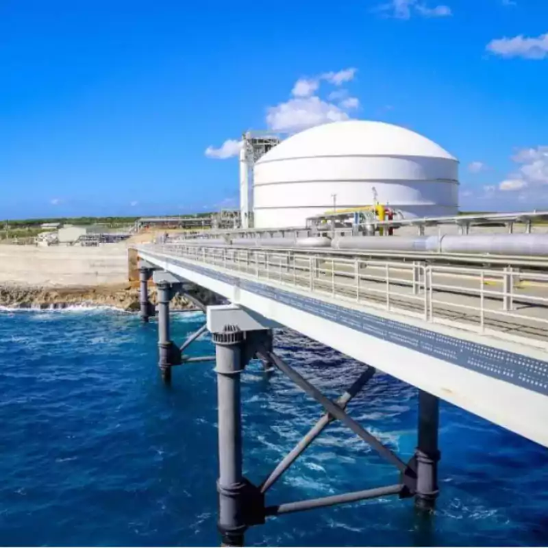 Large white industrial storage tank near the coast with a metal pipeline bridge extending over blue ocean water under a clear sky.