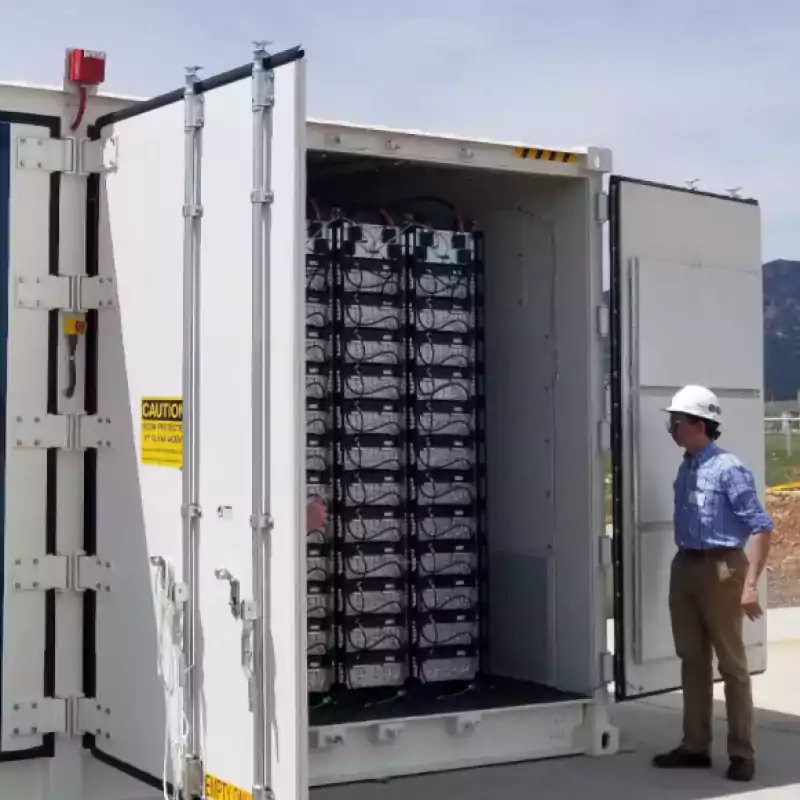 A man wearing a hard hat stands next to an open container filled with stacked industrial batteries outdoors. The container doors display caution signage, and mountains are visible in the background.