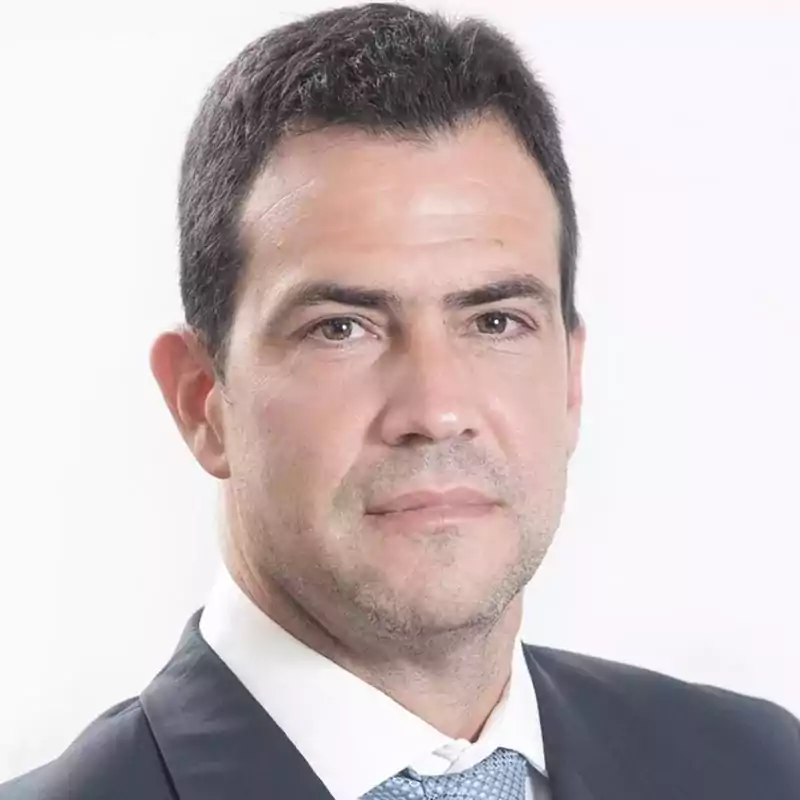 Portrait of a man with short dark hair wearing a dark suit, white shirt, and light blue tie, looking directly at the camera with a neutral expression against a plain white background.