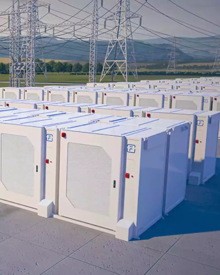 Rows of large white battery storage units are installed outdoors near high-voltage power lines and electrical towers, with a green landscape and hills in the background.