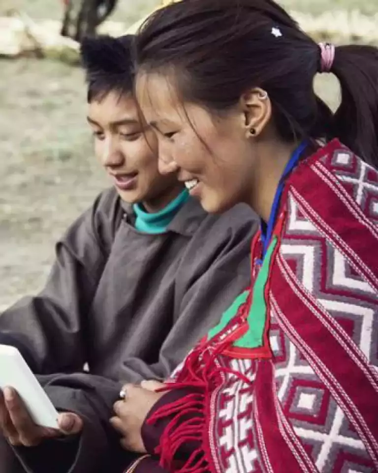 Two people sitting outdoors, smiling and looking at a digital tablet together. One person wears a patterned red shawl, and both appear engaged and happy.