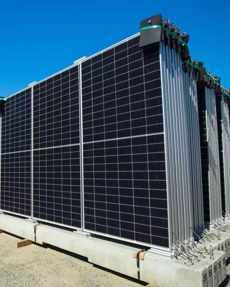 Stacks of large solar panels are arranged outdoors on concrete bases under a clear blue sky, ready for installation at a solar energy site.