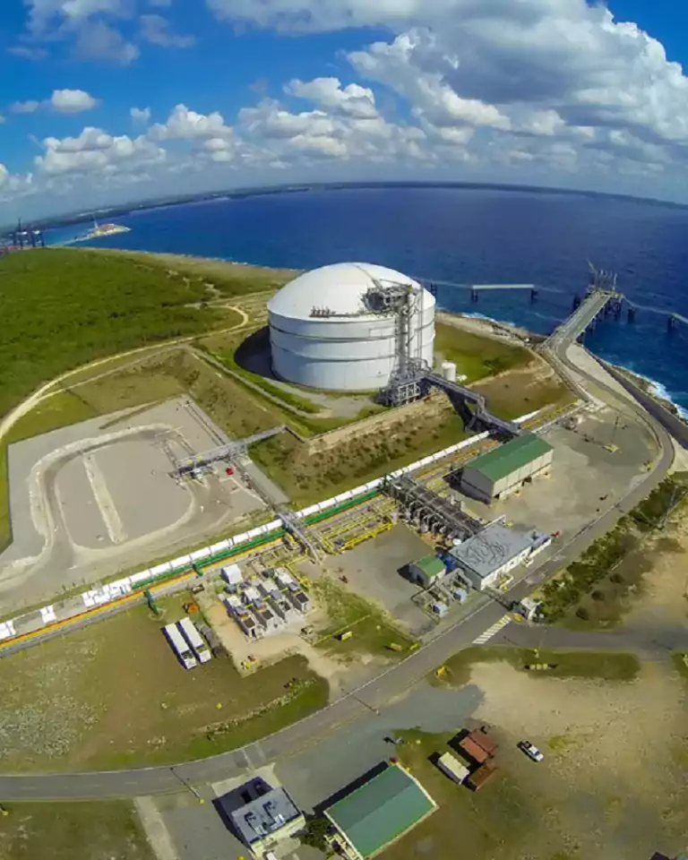 Aerial view of a coastal industrial facility with a large white storage tank, several buildings, roads, and a pier extending into the blue ocean, surrounded by green forest and partly cloudy sky.