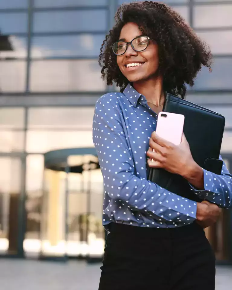 Smiling woman with curly hair and glasses, wearing a blue polka dot shirt, holding a black folder and pink smartphone, standing outside a modern glass office building.