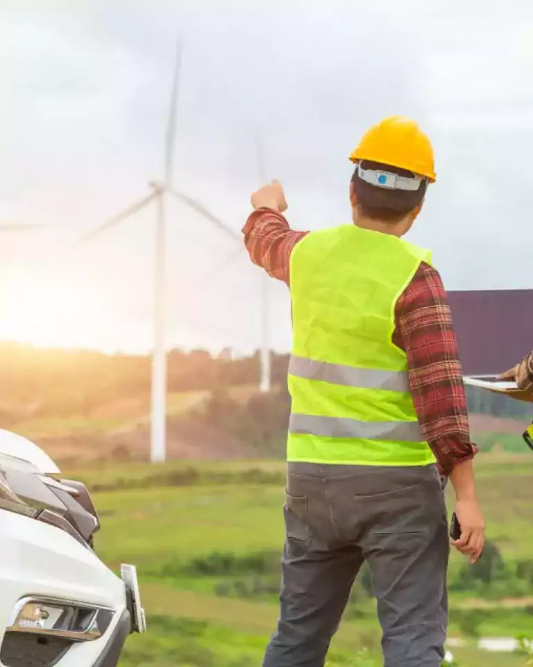 Two engineers wearing safety vests and helmets stand near a white vehicle in a wind farm, with one pointing towards wind turbines and the other holding a laptop, discussing renewable energy operations.