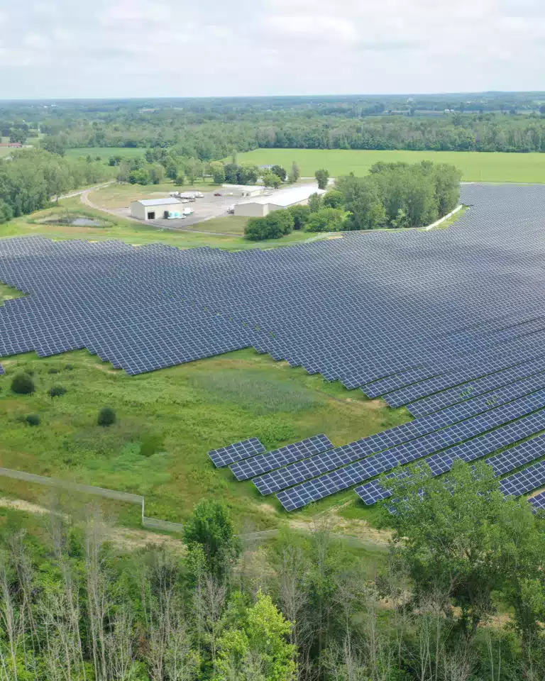 A large solar farm with hundreds of solar panels is surrounded by green fields and trees, with a few buildings in the background under a partly cloudy sky.