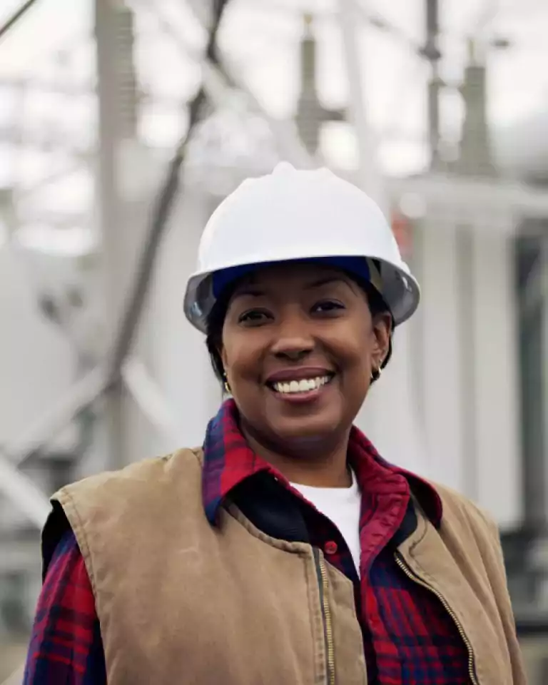 Smiling woman wearing a white hard hat and plaid shirt stands outdoors at an industrial site, representing a construction or engineering worker.