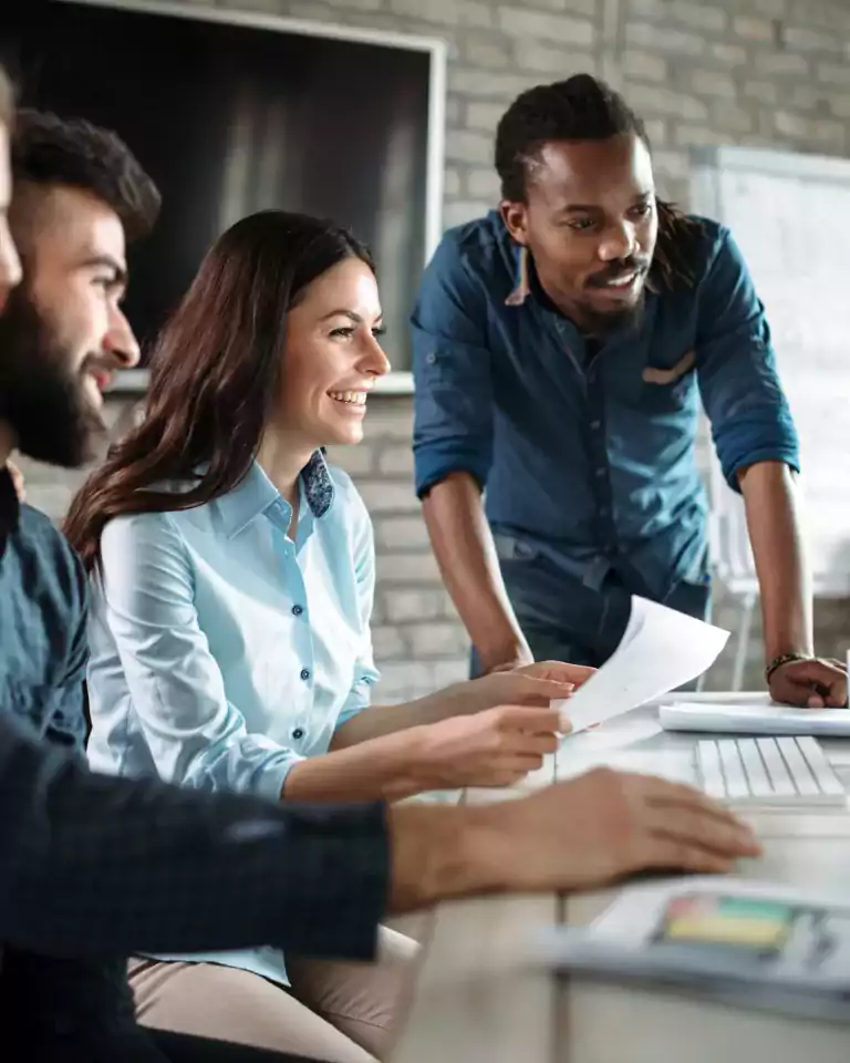 Four young professionals collaborate at a desk in a modern office, smiling and looking at a computer screen together. Papers, a keyboard, and a coffee mug are on the desk, with a whiteboard and monitor in the background.