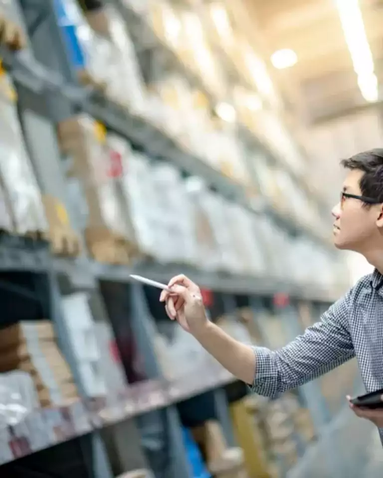 A man wearing glasses and a checkered shirt inspects inventory on tall warehouse shelves, holding a digital tablet and a pen, with boxes and packages organized on the shelves under bright lighting.