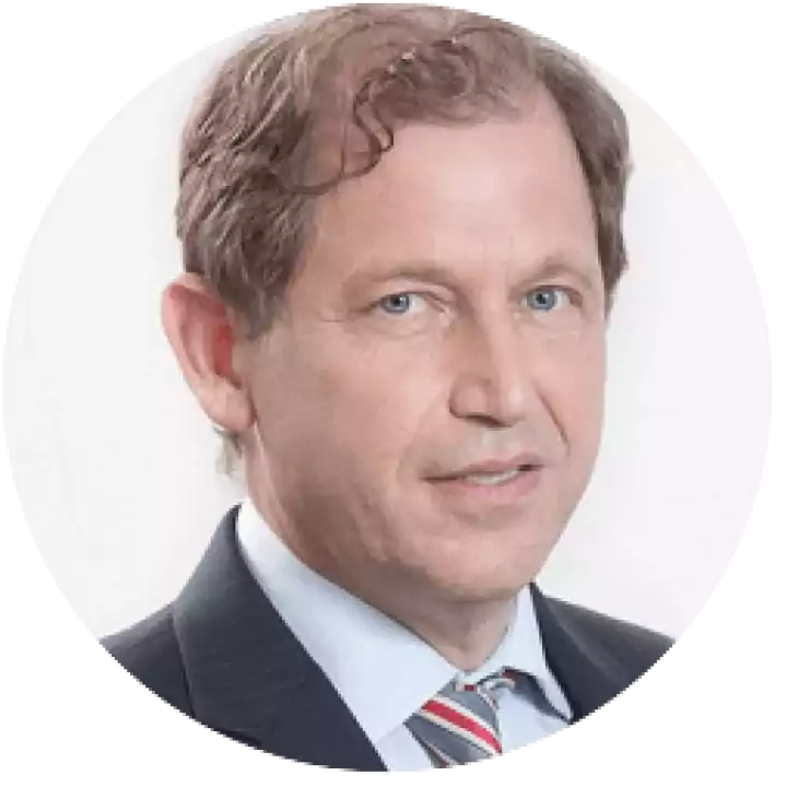 Portrait of a middle-aged man with light brown hair, wearing a dark suit, light blue shirt, and striped tie, looking at the camera with a neutral expression against a white background.