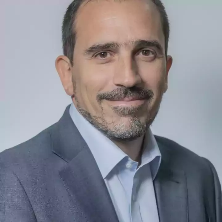 Portrait of a middle-aged man with short dark hair, beard and mustache, wearing a light blue shirt and a gray suit jacket, smiling softly against a plain light background.