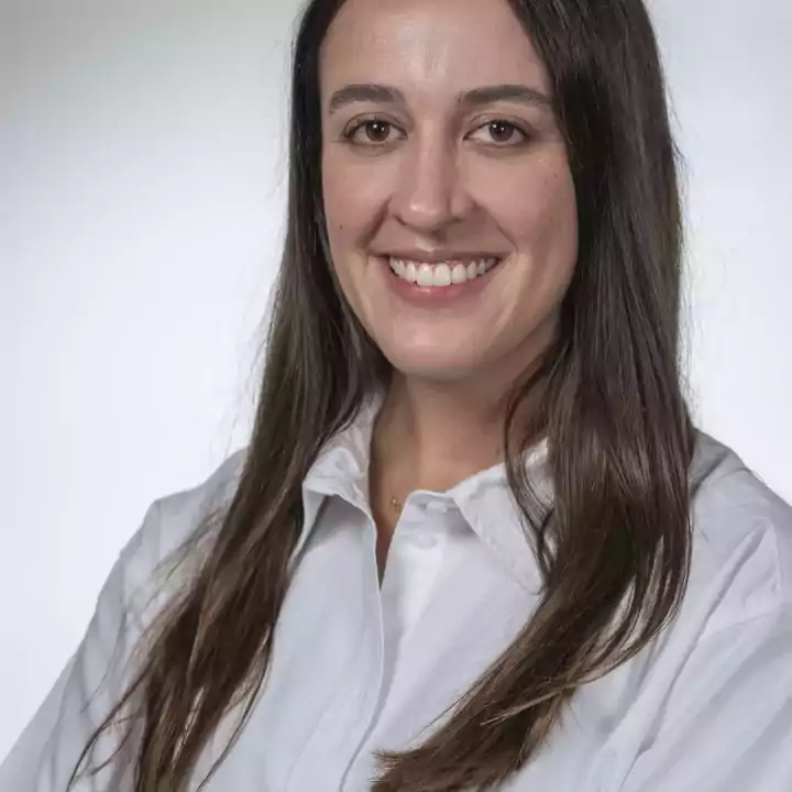 Smiling woman with long brown hair wearing a white button-up shirt, standing with arms crossed against a plain light background.