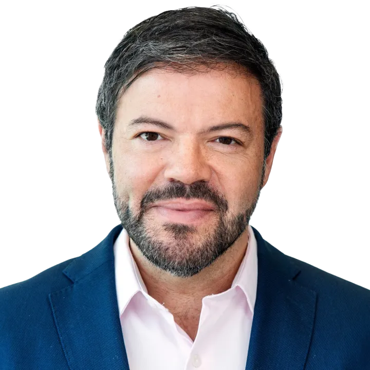 A man with short dark hair and a beard wearing a blue blazer and white shirt, smiling slightly against a plain background.