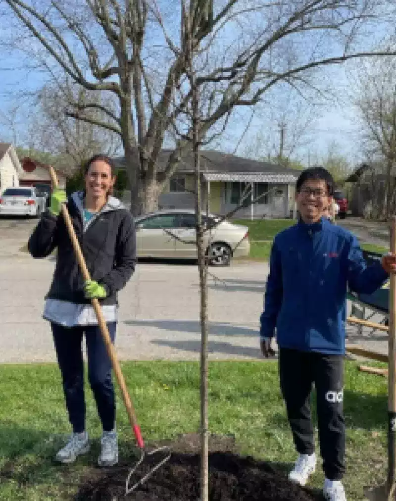 Two people stand outdoors on a grassy area, smiling while holding gardening tools next to a newly planted young tree. Houses, cars, and leafless trees are visible in the background on a clear day.