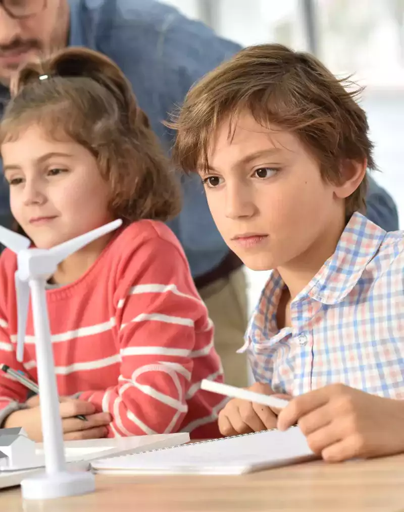 Children in a classroom closely observe a model of wind turbines and houses on a table, guided by an adult, illustrating a renewable energy or science lesson.
