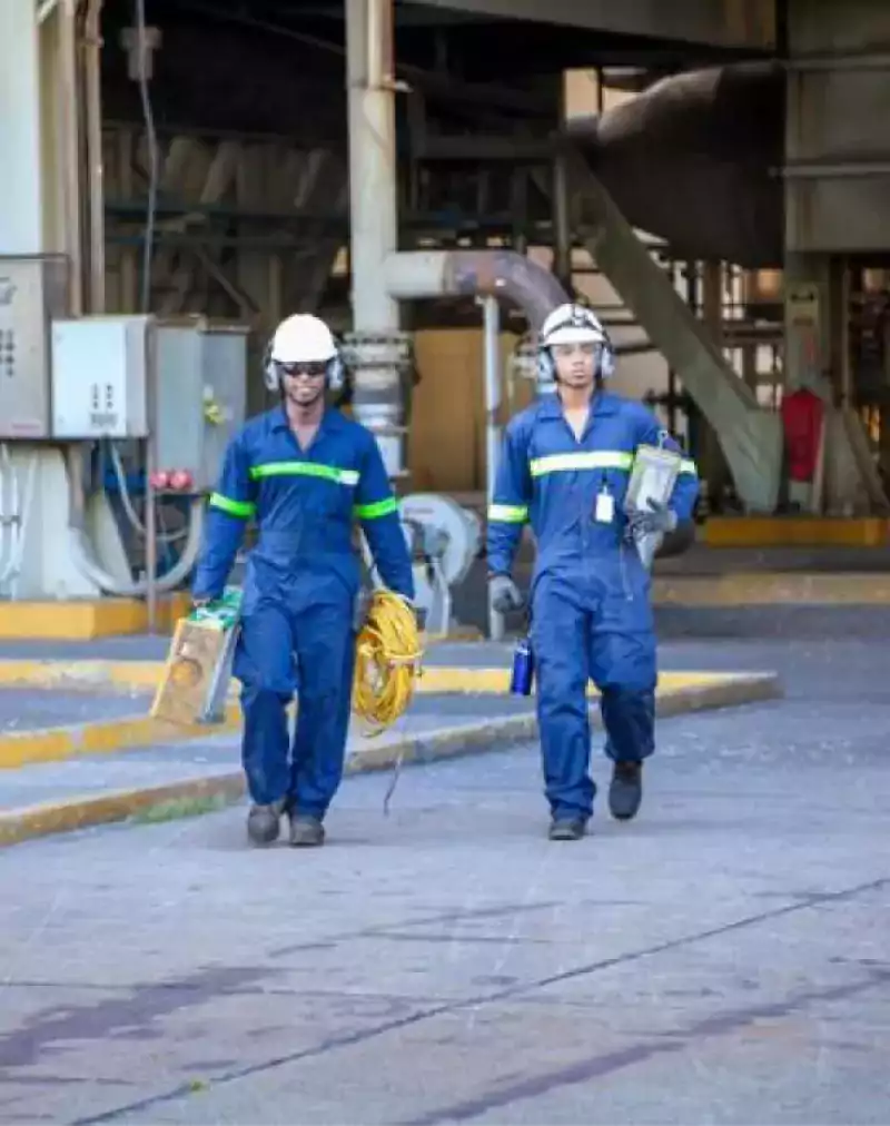 Two industrial workers wearing blue coveralls, safety helmets, and reflective stripes walk in a factory setting, carrying tools and equipment including a yellow cable and a metal box.
