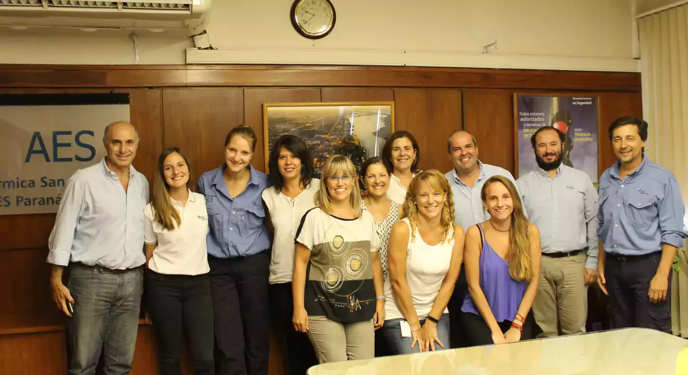 Group of twelve people standing and smiling in an office meeting room with a wooden wall, a clock, and posters in the background. Some are wearing business casual clothes and others are in uniforms. A table with cups and saucers is visible in the foreground.