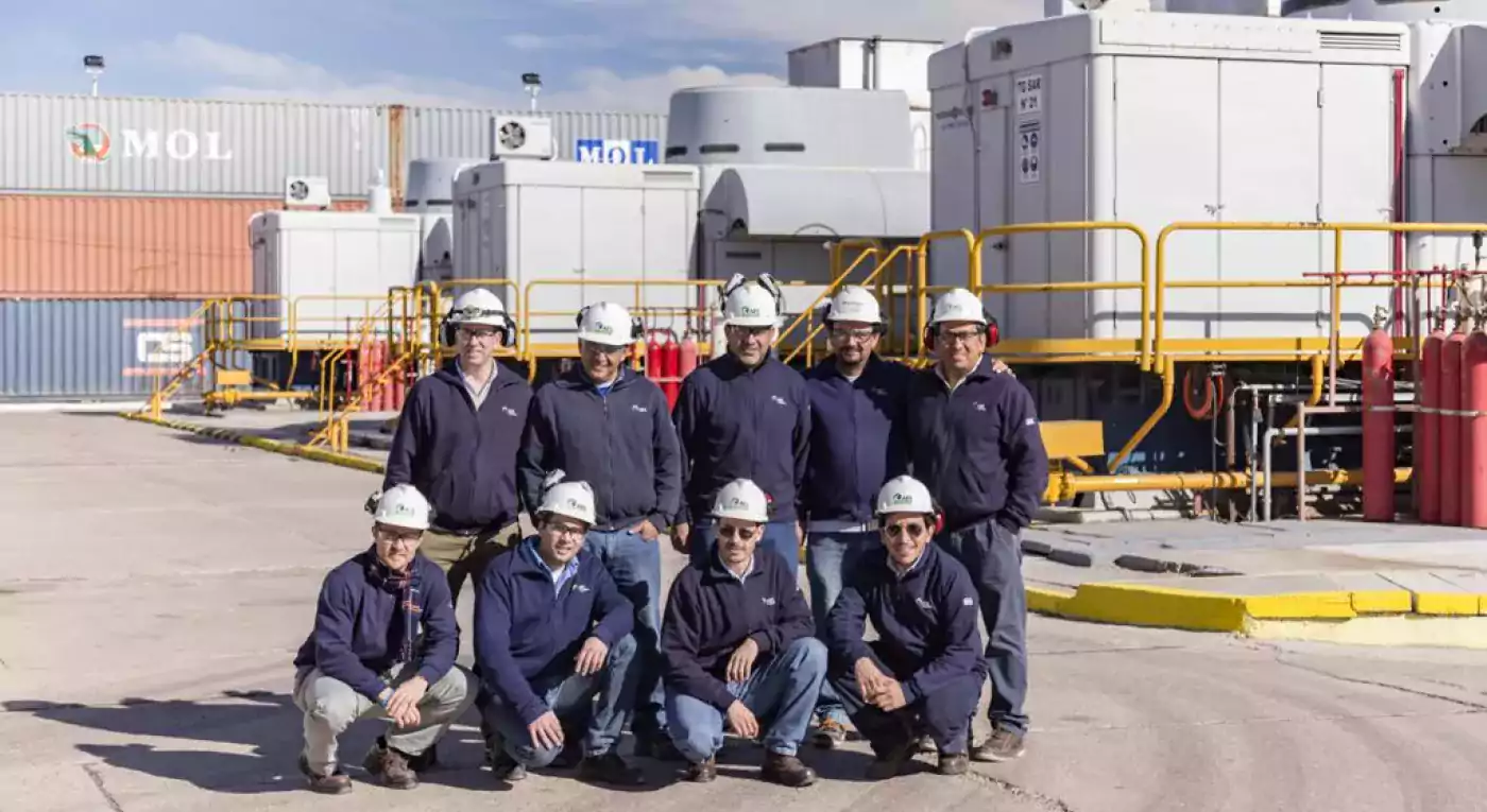 Group of industrial workers wearing navy blue uniforms and white safety helmets posing together outdoors at an industrial facility with containers and machinery in the background.