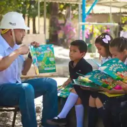 An adult wearing a white hard hat reads a colorful book to three young children sitting outdoors. The children, dressed in school uniforms, are attentively looking at their own copies of the book. Trees and a playground structure are visible in the background.