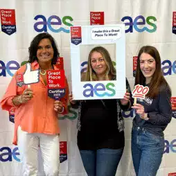 Three women smiling and holding celebratory signs in front of a backdrop with the AES logo and 'Great Place to Work Certified 2023' banners. One holds a frame reading 'I make this a Great Place To Work!' and others hold props showing company pride.