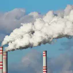 Three industrial smokestacks emitting thick white smoke into the sky, with clouds in the background, illustrating air pollution.