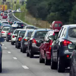 Long line of cars stuck in heavy traffic on a multi-lane road surrounded by trees, with brake lights illuminated and vehicles closely packed.