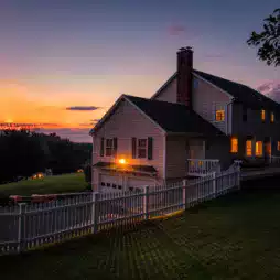 Two-story suburban house with white picket fence and glowing lights, set on a grassy lawn at sunset with a colorful sky in the background.