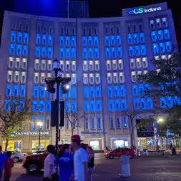 Night view of the AES Indiana building illuminated with blue lights, featuring Old National Bank on the ground floor. Several people walk along the street in front of the building, and cars are parked nearby.