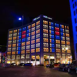 A modern office building at night with the sign 'AES Indiana' on top, illuminated by vertical blue, red, and orange lights. The building is surrounded by parked cars and street lamps, creating a vibrant urban atmosphere.